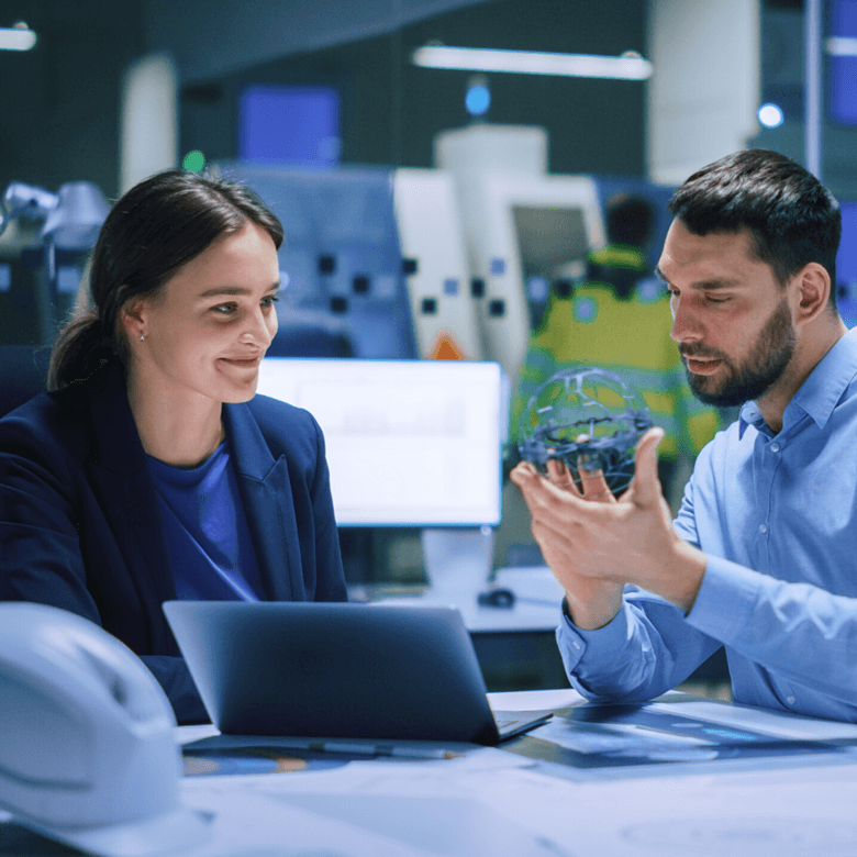 © Adobestock Gorodenkoff Industry 4.0 Modern Factory Meeting Room Chief Engineer Holds Mechanism, Shows It To Female Designer, Use Laptop. Scientists In Contemporary Lab Build Electronic Machinery